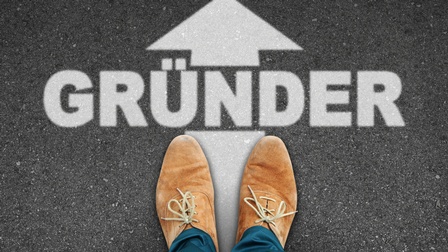 Top-Shot der hellbraunen Schuhe einer Person, die auf einem asphaltierten Boden stehen. Vor den Schuhen steht in weißer Schrift Gründer. Darüber ist ein Pfeil, der in den oberen Bildbereich zeigt