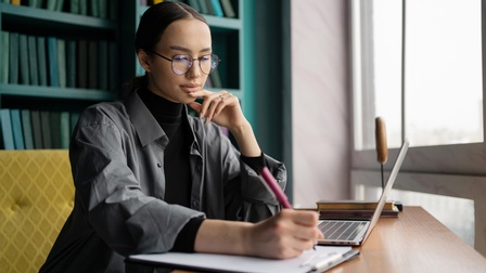 Person mit Brillen sitzt an Tisch vor aufgeklapptem Notebook und nimmt auf Clipboard Notizen vor, im Hintergrund Bücherregal mit vorwiegend grünpetrolfarbenen Buchrücken