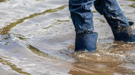 Person in Jeans geht mit blauen Gummistiefeln in tiefem Wasser, Hochwasser