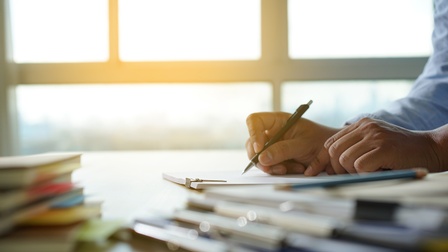Seitliche Aufnahme einer Person, die vor sich viele Unterlagen hat und mit einem Stift etwas darauf unterstreicht