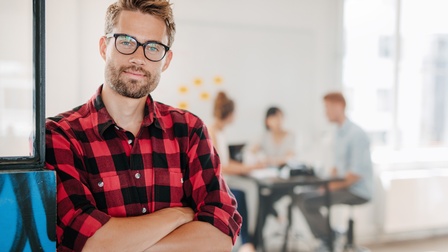 Sanft lächelnde Person mit verschränkten Armen in rot-schwarz kariertem Hemd im Fokus, im Hintergrund Personen an Tisch sitzend