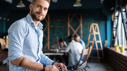 Person in blau-weiß gestreiften Hemd lächelt und hat die Hände auf der Laptoptastatur, im Hintergrund weitere Personen am Tisch sitzend