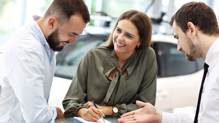Drei lächelnde Personen, eine Hält Stift auf Dokument, im Hintergrund Auto