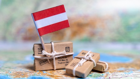Nahaufnahme einer Landkarte, auf der drei kleine Miniaturpäckchen liegen, auf denen Adressen stehen. Um jedes Päckchen ist ein Band gewickelt. In einem Band steckt die Flagge Österreichs. Der Hintergrund bildet verschwommen ebenfalls eine Landkarte ab. 