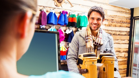 Porträt einer Person mit Jacke und Schale, die in einem Geschäft an der Kassa steht. Vor der Person stehen zwei Skischuhe. Hinter der Kassa steht eine Person. An der Wand hängen Mützen