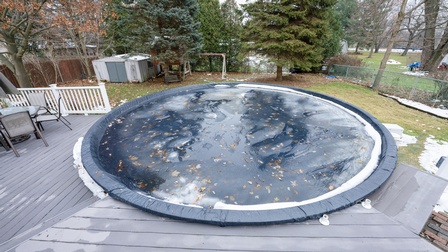 Topshot eines runden Pools im Garten, der in eine Veranda aus schwarzem Holz eingefasst ist. Der Pool ist mit einer Plane abgedeckt, auf der sich Wasser und Laub sammeln. Es ist ein nebeliger Herbsttag. 