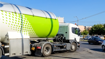 Ein Lastkraftwagen mit einem zylindrischen Behälter steht schräg auf einer asphaltierten Straße, die auf eine weitere asphaltierte Straße trifft, wo Autos sind.