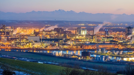 In der Bildmitte sind verschiedene Gebäude. Zwischen den Gebäuden stehen Rohre und Silos. Aus manchen Gebäuden und Rohren steigt Rauch auf. Vor manchen Gebäuden ist ein Gewässer. Im Vordergrund ist eine Wiese