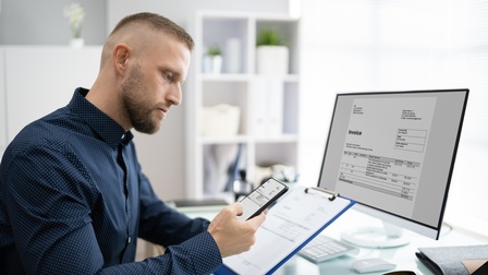 Person mit blau gepunktetem Hemd sitzt mit einem Smartphone und einem Klemmbrett vor einem Schreibtisch mit Bildschirm und betrachtet Rechnungen