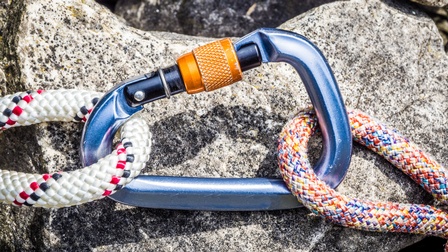 Nahaufnahme eines Karabiners, der auf einem Stein liegt und bei dem links und rechts ein Seil eingehängt ist