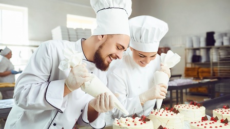 Zwei junge lächelnde Patisseur:innen in Arbeitskleidung beim Verzieren von Torten