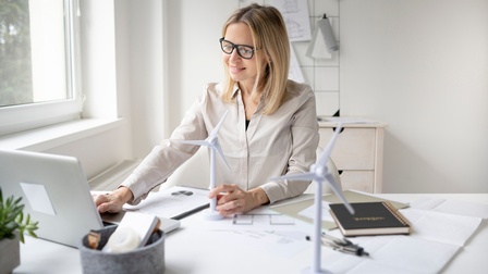Person mit Brille sitzt an Arbeitstisch, blickt auf aufgeklappten Laptop vor sich und hält Modell eines Windrades in der Hand, ringsum am Tisch Arbeitsunterlagen, Pläne, ein weitere Windradmodell und ein Zirkel