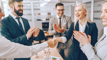 Zwei Personen geben sich die Hand in einem Besprechungsraum während andere Personen sie wohlwollend und lächelnd anblicken und beifallend klatschen