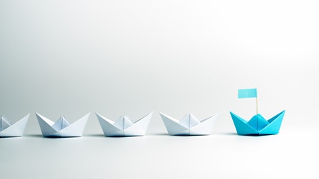 Mehrere weiße Papierschiffchen nebeneinander platziert, an vorderster Stelle ein türkises Papierschiffchen mit Fahne