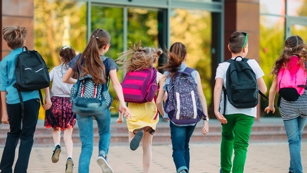 Rückenansicht mehrerer Kinder mit Rucksäcken, die in Richtung eines Gebäudeeingangs laufen.