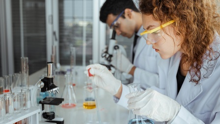 Person mit Schutzbrillen in weißem Kittel an Tisch sitzend, mit Pipette Flüssigkeit in Schälchen tropfend hält in Hand Glas mit Flüssigkeit, am Tisch Mikroskop und Röhrchen mit Flüssigkeiten, im Hintergrund weitere Person durch Mikroskop äugend
