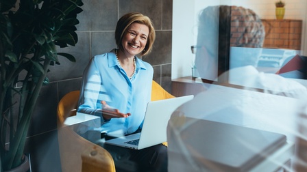Lächelnde Person sitzt in gelbem Stuhl mit aufgeklappten Notebook am Schoß und unterhält sich mit anderer Person in Rückenansicht