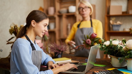 Lächelnde Person mit Schürze sitzt vor aufgeklapptem Notebook, ringsum Blumen, im Hintergrund weitere Person mit Schürze stutzt Blumenstrauß
