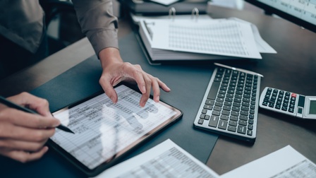 Eine Person tippt auf einem Tablet, auf dem eine Tabelle zu sehen ist. Es sind nur ihre Hände zu sehen. Das Tablet liegt auf einem Schreibtisch, auf dem Unterlagen und ein Taschenrechner liegen. 