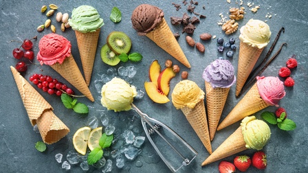 Verschiedene Waffeltüten mit Eiskugeln auf dunklem Hintergrund, dazwischen Erdbeeren, Nüsse, Schokoladestücke, Kiwis, Zitronenscheiben, Nektarinen, Himbeeren, Vanilleschoten, Eiswürfel, Minzblätter etc. Mittig ist ein Eisportionierer mit einer Eiskugel 