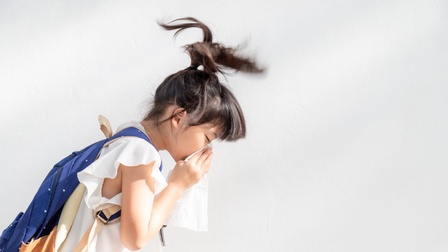 Seitliche Aufnahme eines Kindes mit Rucksack, das vor einer hellen Wand steht und sich in ein Taschentuch schnäuzt