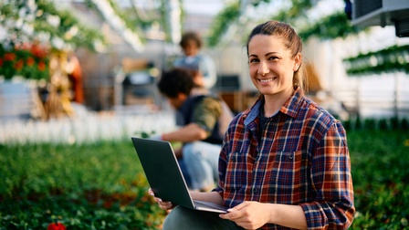 Nahaufnahme einer Person, die mit aufgeklapptem Laptop auf einer Grasfläche sitzt. Im Hintergrund ist verschwommen eine Person zu erkennen, die im Gras Gartenarbeit verrichtet , ebenso ein Gewächshaus