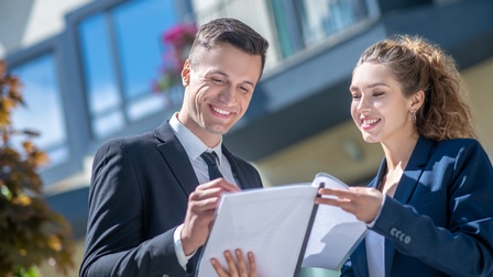 Zwei Personen in dunkelblauen Sakkos stehen nebeneinander im Freien vor einem Haus. Beide lächeln. Die rechte Person hält der linken Person einen Vertrag hin, den die linke Person anblickt