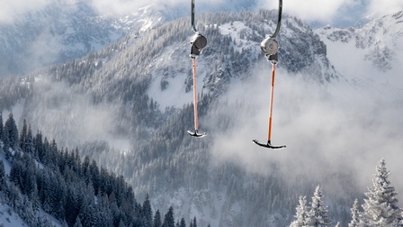 Zwei schwarze, kurze, horizontale Stangen sind mit einer langen orangen Stange verbunden, die wiederum an einem Seil hängen. Im Hintergrund sind schneebedeckte Hänge mit schneebedeckten Wäldern