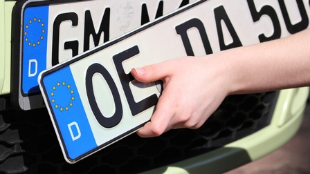 Hand hält ein weiteres Autokennzeichen zu einem Fahrzeug mit montiertem Autokennzeichen