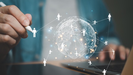 Person hält einen weißen Stift in der Hand auf dem sich eine Weltkugel mit Personen-Icons, die vernetzt sind in der Farbe weiß visualisieren, daneben steht ein Laptop auf einem Tisch