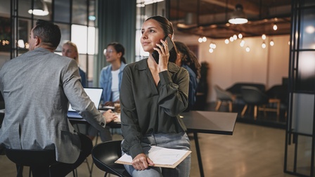 Person an hohem Stuhl lehnend hält Smartphone an Ohr und blickt zur Seite, am Schoß hält sie einen Block, im Hintergrund Tisch mit weiteren Personen rundherum sitzend<br />
