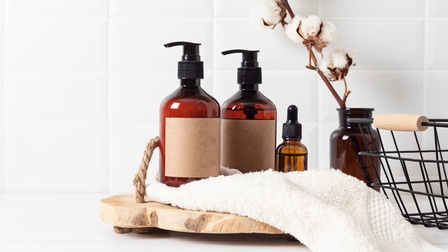 Pflegeprodukte stehen auf einem Holzuntergrund, dahinter eine Vase mit Bambusknospen, daneben auch ein Drahtkorb, vor einer weißen Kulisse