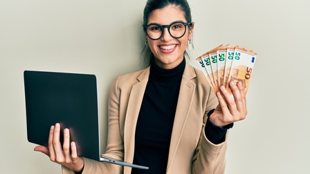 Halbporträt einer lächelnden Person in einem schwarzen Oberteil mit Sakko und Brillen, die vor einer hellen Wand steht. In der rechten Hand hält sie einen aufgeklappten Laptop. In der linken Hand hält sie mehrere Fünfzig-Eurogeldscheine.