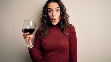 Eine Person in einem dunkelroten Rollkragenpullover, mit langen, dunkelbraunen Locken steht vor hellgrauem Hintergrund. In der rechten Hand hält sie ein Glas Wein befüllt mit Rotwein. Ihr Mund ist zu einem O geformt