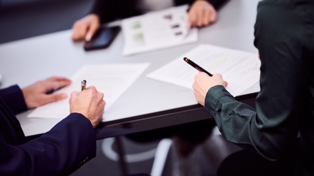 Over the shoulder-Shot von Personen an einem Tisch, die ein Dokument unterschrieben, sie halten in ihrer rechten und ihrer linken Hand einen Stift. Eine Person sitzt ihnen gegenüber und blättert in einem Papierstapel. 