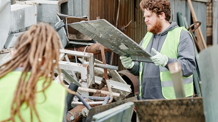 Zwei Personen in gelben Warnwesten beim Sortieren von Altmetallen, eine Person mit Handschuhen und Bart im Fokus, die großen Metalldeckel in Händen hält, die andere Person in Rückenansicht, ringsum teils rostige Metallgegenstände