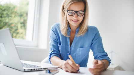 Sitzende Person mit langen blonden Haaren und Brille in blauem Hemdmacht Notizen, daneben Laptop aufgeklappt und Smartphone liegend