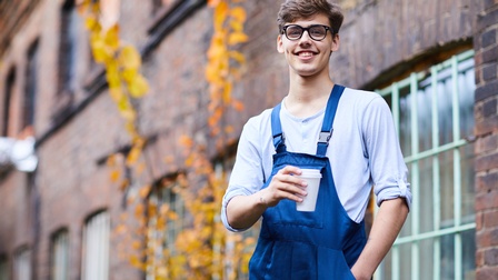 Person mit kurzen dunklen Haaren, Brille und blauer Latzhose steht freudig und blickt in die Kamera während ein Kaffeebecher gehalten wird, im Hintergrund zeigt sich ein Gebäude mit Backsteinen