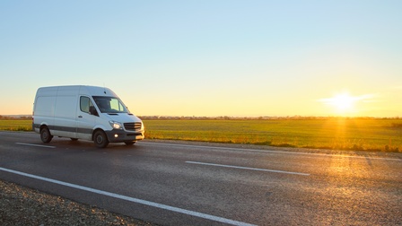 Ein weißer Kleinlastwagen fährt auf einer zweispurigen Straße auf der linken Fahrspur. Neben der Straße ist Wiese. Am Horizont steht die Sonne tief