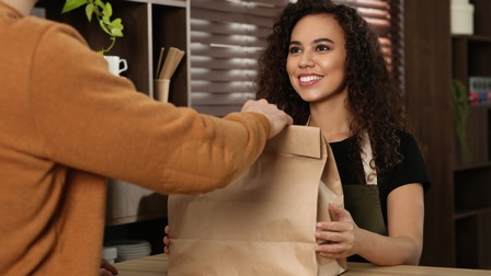 Zwei Personen stehen sich an einem Tresen gegenüber. Die rechte Person lächelt, trägt eine Schürze und reicht der anderen Person eine braune Einkaufstasche