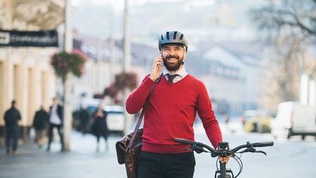 Frontale Aufnahme einer Person mit rotem Pulli, Krawatte, dunkler Hose, Helm und Tasche schiebt mit der linken Hand ein Fahrrad. Mit der rechten Hand telefoniert die Person und lächelt. Im Hintergrund sind Gebäude und Leute