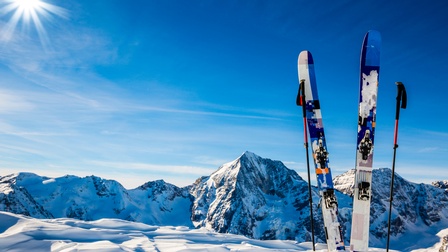 Zwei Ski stecken vertikal in einer Schneedecke. Links und rechts davon sind jeweils ein Skistock. Im Hintergrund sind schneebedeckte Berge. Der Himmel ist blau, man sieht in der linken Bildhälfte das Licht der Sonne. 