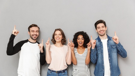 Vier Personen in Jeans und T-Shirts stehen nebeneinander vor einer grauen Wand und lächeln. Jede Person blickt in die Kamera und deutet mit beiden Zeigefingern jeweils in die Luft