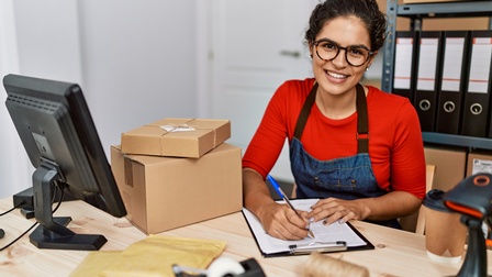 Eine lächelnde Person sitzt im Büro und schreibt auf einem Zettel, welches am Clipboard hängt. Auf dem Tisch stehen ein Computer, Pakete, ein Scanner und Klebeband. Im Hintergrund ist ein Regal mit Mappen zu sehen.