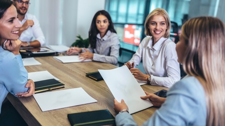 Um einen Tisch sitzen fünf Personen. Vor jeder Person sind Zettel und ein Notizbuch. Alle blicken zu einer Person, die an einem Tischende sitzt