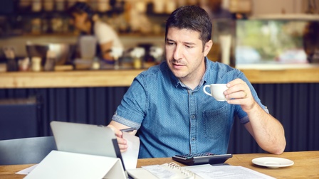 Person sitzt in einem Lokal und blickt auf sein Tablet und hält dabei eine Tasse in der Hand, am Tisch liegt ein Kalender, sowie Unterlagen und ein Rechner, im Hintergrund sieht man die Theke sowie eine mitarbeitende Person arbeiten