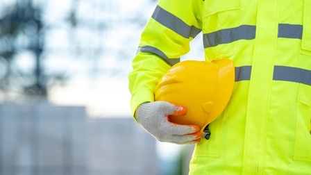 In der rechten Bildhälfte ist der Oberkörper einer Person. Die Person trägt eine neongelbe Jacke und Handschuhe. Mit der rechten Hand hält sie einen gelben Helm