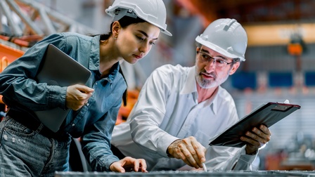 Zwei Personen mit Helmen stehen nebeneinander und lehnen sich mit den Oberkörpern leicht nach vorne über eine Oberfläche, auf die sie beide blicken. Die linke Person hält ein Klemmbrett, die rechte Person ein Tablet.