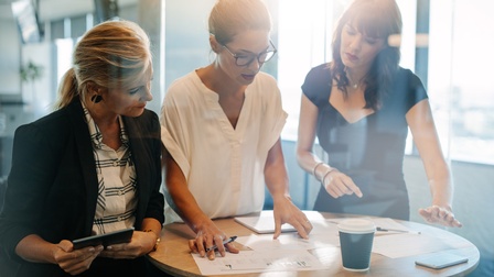 Personen in Businesskleidung stehen rund um einen Stehtisch und begutachten Unterlagen