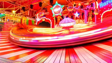 Auf einem Boden mit abwechselnd roten und weißen Brettern ist ein sich schnell drehendes kreisförmiges Rummelgefährt, die Oberfläche des Objekts in der Bewegung verschwommen. An der Wand dahinter sind verschiedene Personen aufgezeichnet.